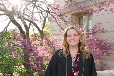 Noel Mills in her graduation gown standng in front of flowering tree on the University of Iowa campus