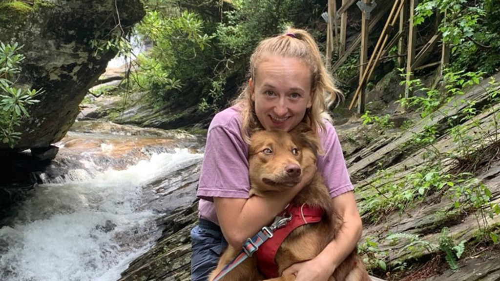 Lily Sronkoski holding her dog with a forest and stream in the background